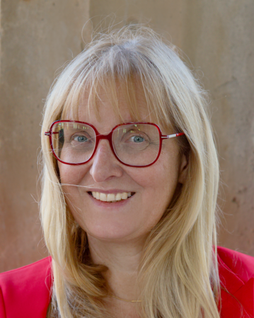 Eine Frau mit langen blonden Haaren und roten Brillenläsern lächelt freundlich. Sie trägt ein rotes Oberteil und steht vor einer neutralen Wand.