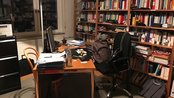 Ein Arbeitszimmer mit einem Schreibtisch, einem Computer, Büchern in Regalen und einem Stuhl. Auf dem Tisch liegen Unterlagen und eine Wasserflasche. Im Hintergrund sind ein Aktenschrank und eine Mülltonne sichtbar.