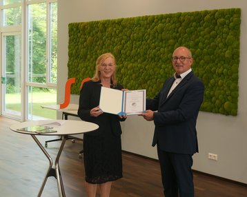 Zwei Personen stehen in einem modernen Raum. Eine Frau überreicht einem Mann ein Dokument. Im Hintergrund ist eine grüne Mooswand zu sehen.