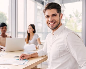 Ein lächelnder Mann sitzt an einem Tisch in einem Besprechungsraum, umgeben von drei weiteren Personen. Auf dem Tisch liegen Dokumente und ein Laptop.