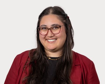 Eine lächelnde Frau mit langen, dunklen Haaren trägt eine rote Jacke und eine schwarze Bluse. Sie hat eine Brille und steht vor einem neutralen Hintergrund.