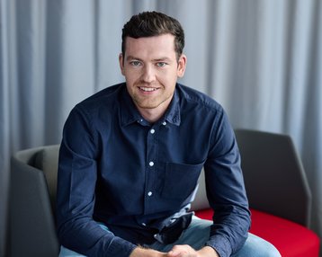 Ein junger Mann mit dunklem Hemd sitzt auf einem Stuhl und lächelt, während er die Hände auf den Knien hält.