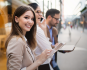Eine lächelnde Frau in einem Blazer hält ein Tablet, während sie mit zwei anderen Personen in einem urbanen Umfeld steht. Die Gruppe scheint an einem geschäftlichen Austausch interessiert zu sein.