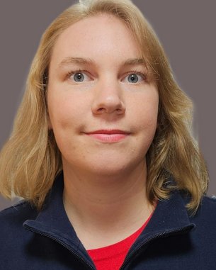 Eine Person mit blonden, schulterlangen Haaren und blauen Augen trägt einen dunkelblauen Pullover. Der Hintergrund ist einfarbig und neutral.