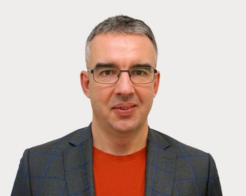 Mann mit Brille, grauen Haaren und einem grauen Blazer mit Karomuster, trägt ein rotes T-Shirt. Neutraler Hintergrund.