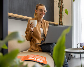 Eine lächelnde Frau sitzt auf einem Sofa, hält eine Tasse in der Hand und arbeitet an einem Laptop. Im Hintergrund sind Pflanzen und ein Tisch mit einer Schüssel Obst zu sehen.
