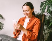Eine Frau sitzt entspannt in einem Stuhl und schaut auf ihr Smartphone. Sie trägt ein orangefarbenes Hemd und graue Jeans, umgeben von Pflanzen im Hintergrund.