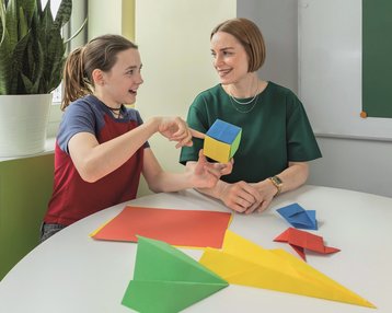 Eine Lehrerin und ein Schüler sitzen an einem Tisch und diskutieren über bunte Papierformen, während sie eine geometrische Figur halten.