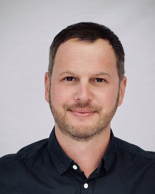 Ein Mann mit kurzem, dunklem Haar und einem leichten Bart lächelt in die Kamera. Er trägt ein dunkelblaues Hemd und hat einen neutralen Gesichtsausdruck vor einem hellen Hintergrund.
