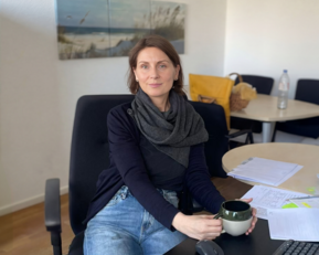 Frau mit einem schwarzen Schal sitzt an einem Schreibtisch und hält eine Tasse in der Hand, umgeben von Unterlagen und Büromaterial.