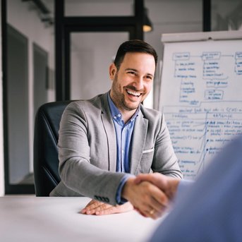 Ein Mann in einem grauen Anzug schüttelt einem anderen Mann die Hand. Im Hintergrund steht ein Flipchart mit Notizen.