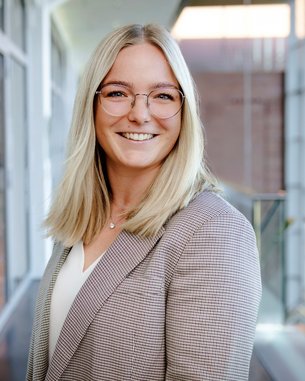 Eine lächelnde Frau mit blonden Haaren und Brille trägt einen karierten Blazer. Sie steht in einem modernen Innenraum mit großen Fenstern.