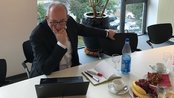 Ein Mann in einem Anzug sitzt an einem Tisch mit einem Laptop, während er nachdenklich auf den Bildschirm schaut. Auf dem Tisch befinden sich Getränke und Snacks, darunter Obst und Gebäck.