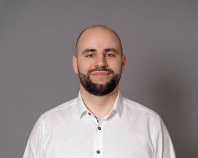 Ein Mann mit kurzem Haar und Bart trägt ein weißes Hemd. Er steht vor einem grauen Hintergrund und lächelt leicht.