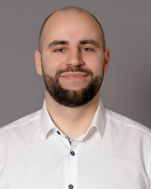 Ein Mann mit kurzem Haar und Bart trägt ein weißes Hemd. Er steht vor einem grauen Hintergrund und lächelt leicht.