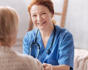 Eine Krankenschwester in blauer Uniform lächelt, während sie die Hand einer älteren Patientin hält. Ein Stethoskop hängt um ihren Hals.