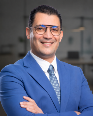 Ein Mann in einem blauen Anzug mit Brille lächelt und steht mit verschränkten Armen in einem modernen Büro.