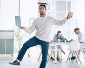 Ein Mann balanciert ein Buch auf dem Kopf, während er einen Laptop in einer Hand und einen Kaffeebecher in der anderen hält.