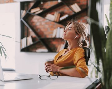Eine Frau mit Kopfhörern sitzt entspannt an einem Schreibtisch, hält einen Kaffeebecher in der Hand und blickt nach oben. Im Hintergrund sind Bücherregale und Pflanzen sichtbar.