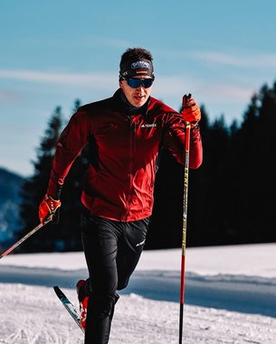 Ein Skilangläufer in roter Jacke und schwarzer Hose bewegt sich auf einer verschneiten Strecke, umgeben von Bäumen und Bergen im Hintergrund.