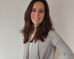 Eine Frau mit langen, braunen Haaren trägt einen grauen Blazer und lächelt. Der Hintergrund ist neutral und hell.