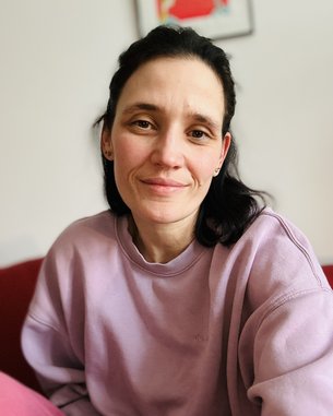 Eine Person mit dunklen Haaren trägt einen lila Pullover und sitzt auf einem roten Sofa, lächelnd in die Kamera.