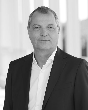 Ein Mann in einem schwarzen Anzug steht vor einer hellen, modernen Architektur. Er hat kurzes, graues Haar und lächelt leicht.