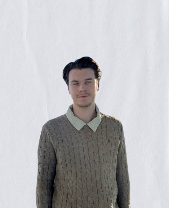 Junger Mann mit dunklen Haaren, der einen beigen Pullover mit Zopfmuster und einem hellen Kragen trägt. Er steht vor einem neutralen Hintergrund und schaut direkt in die Kamera.