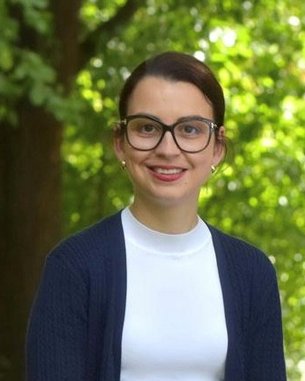 Eine Person mit Brille und einem blauen Cardigan lächelt vor einer grünen, bewaldeten Kulisse.
