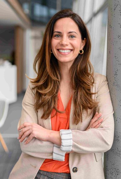 Eine lächelnde Frau mit langen Haaren steht an einer Säule in einem modernen Büro, gekleidet in einem beigen Blazer und orangefarbenem Oberteil.