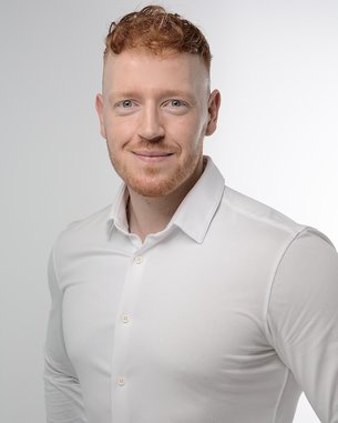 Mann mit roten Haaren und Bart, trägt ein weißes Hemd, lächelt in die Kamera vor einem hellen Hintergrund.