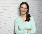 Eine Frau mit Brille und langen, braunen Haaren steht vor einer weißen Ziegelwand. Sie trägt ein hellblaues Oberteil und lächelt, während sie die Arme vor der Brust verschränkt.