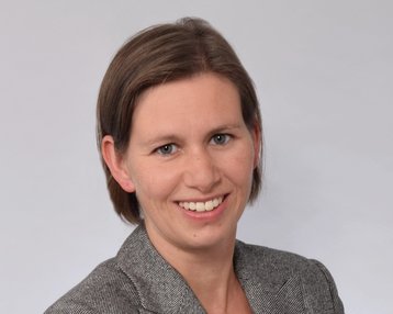 Eine Frau mit kurzen, braunen Haaren lächelt und steht mit verschränkten Armen. Sie trägt einen grauen Blazer über einem weißen Oberteil. Der Hintergrund ist hell und neutral.