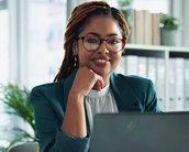 Eine Frau mit lockigen, geflochtenen Haaren und Brille sitzt an einem Schreibtisch, lächelt und schaut auf einen Laptop.