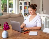 Eine Frau sitzt an einem Tisch und arbeitet an einem Laptop. Vor ihr liegt ein Notizblock und ein roter Stift, während eine Vase mit rosa Blumen auf dem Tisch steht.