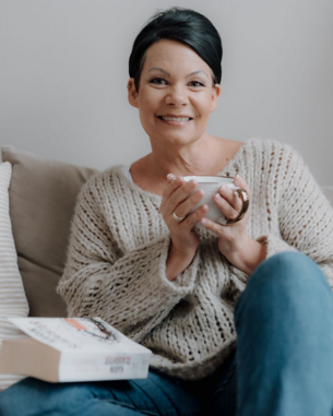 Eine Frau mit kurzem, dunklem Haar sitzt auf einem Sofa, hält eine Tasse in der Hand und lächelt. Neben ihr liegt ein Buch auf dem Sofa. Sie trägt einen gemütlichen, gestrickten Pullover und Jeans.