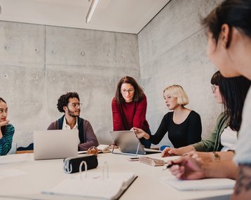 Eine Gruppe von sechs Personen sitzt an einem Tisch in einem modernen Besprechungsraum und diskutiert, während Laptops und Notizen auf dem Tisch liegen.