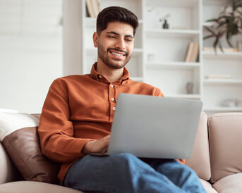 Mann mit Laptop Ein junger Mann sitzt entspannt auf einem Sofa und arbeitet an einem Laptop. Er trägt ein oranges Hemd und lächelt, während er auf den Bildschirm schaut.