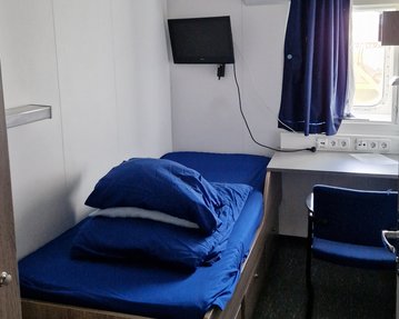 Ein einfaches Zimmer mit einem Einzelbett, blauen Bettwaren, einem Schreibtisch, einem Stuhl und einem Fernseher an der Wand. Ein Fenster mit Vorhängen lässt Tageslicht herein.