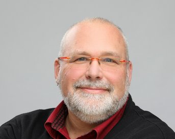 Ein Mann mit Brille und grauem Bart lächelt freundlich. Er trägt ein schwarzes Oberteil mit einem roten Hemd darunter. Der Hintergrund ist neutral und hell.