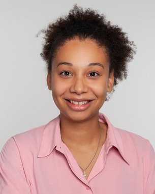 Junge Frau mit lockigem, braunem Haar und einem freundlichen Lächeln, trägt ein rosa Hemd. Der Hintergrund ist neutral und hell.