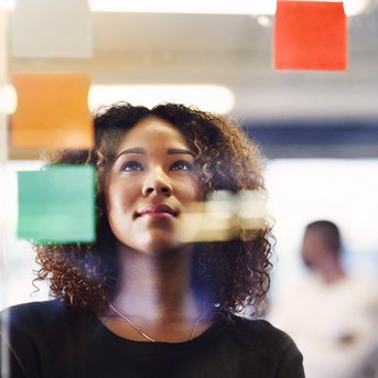 Eine Frau mit lockigem Haar schaut nachdenklich auf bunte Haftnotizen, die an einer Glaswand befestigt sind. Im Hintergrund ist eine weitere Person unscharf zu sehen.