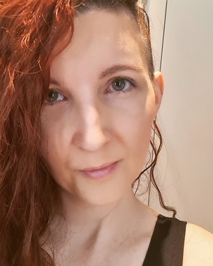Eine Person mit langen, lockigen, roten Haaren schaut direkt in die Kamera. Sie trägt ein schwarzes Oberteil und hat einen neutralen Gesichtsausdruck.