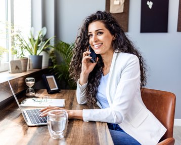 Eine Frau mit lockigem Haar sitzt an einem Schreibtisch, spricht am Telefon und lächelt. Vor ihr steht ein Laptop, ein Glas Wasser und einige Pflanzen sind im Hintergrund sichtbar.