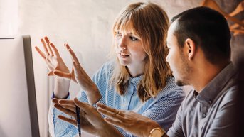 Zwei Personen diskutieren vor einem Computerbildschirm. Die Frau zeigt mit den Händen auf den Bildschirm, während der Mann aufmerksam zuhört und einen Stift in der Hand hält.
