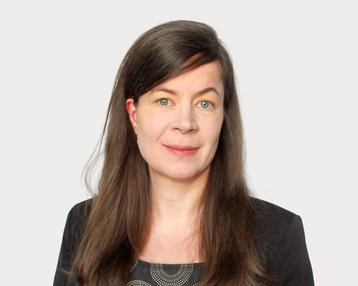 Eine Frau mit langen, braunen Haaren und blauen Augen trägt ein schwarzes Oberteil mit einem grafischen Muster.