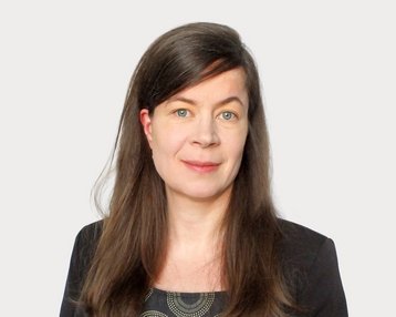 Eine Frau mit langen, braunen Haaren und blauen Augen trägt ein schwarzes Oberteil mit einem grafischen Muster.