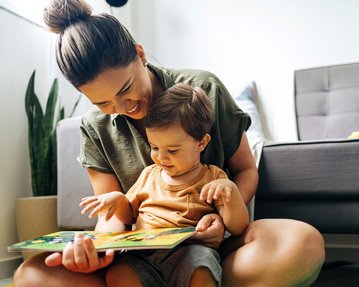 Eine Mutter liest einem kleinen Kind ein Buch vor, während sie auf dem Boden sitzt. Beide lächeln und zeigen Interesse an der Geschichte.