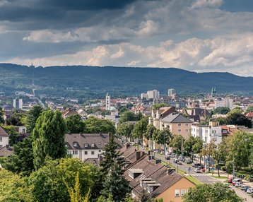 Stadtansicht mit Wohnhäusern, Bäumen und einer Berglandschaft im Hintergrund unter einem bewölkten Himmel.