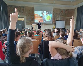 Ein Redner steht vor einer Gruppe von Schülern, die aufmerksam zuhören. Viele Kinder heben die Hände, um Fragen zu stellen. Im Hintergrund ist eine Präsentation auf einem Bildschirm zu sehen.